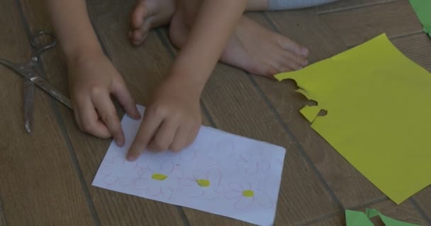 Niña con el pelo rubio, en camiseta rosa y pantalones grises, está sentada en un piso, poniendo medias amarillas a las flores blancas dibujadas — Vídeos de Stock