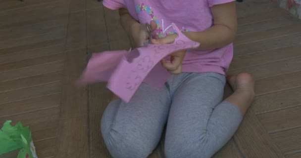 Menina com cabelo loiro, em camiseta rosa e calças cinza, está sentada, cortando flores cor de rosa, colas, papel verde coroa está no chão — Vídeo de Stock