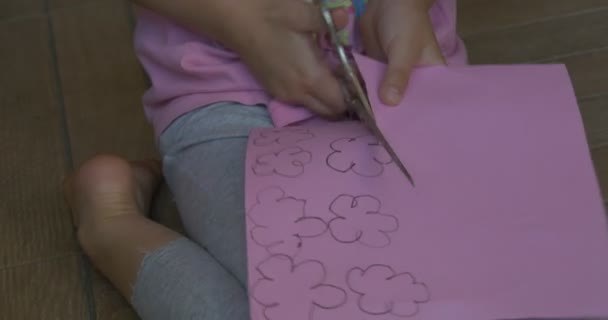 Menina com cabelo loiro, em camiseta rosa e calças cinza, está sentada em um chão, papel de cor, cortando as flores da folha de papel rosa — Vídeo de Stock