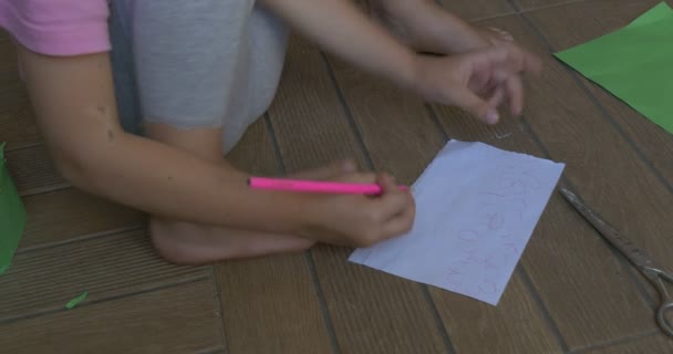 Liten flicka med blont hår, i rosa T-shirt och grå byxor, sitter på ett golv, ritning på det vita arket av papper, grön papper krona — Stockvideo