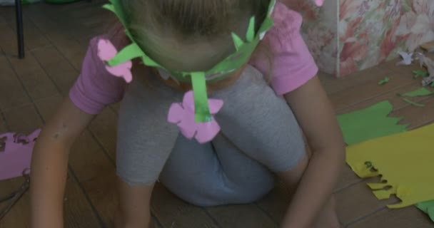 Μικρό κορίτσι με ροζ T-shirt με ξανθιά πλεξούδα κάθεται στο πάτωμα, φτιάχνοντας ένα σχήμα από έγχρωμο χαρτί, καθισμένος σε ένα πράσινο στέμμα, παιδικό βιβλίο — Αρχείο Βίντεο