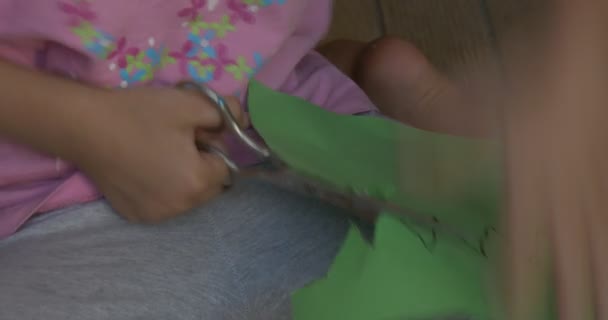 Kleines Mädchen in oinfarbenem T-Shirt sitzt auf dem Boden, Farbpapier vor sich, schneidet die Figur der Krone aus grünem Blatt Papier, Nahaufnahme — Stockvideo