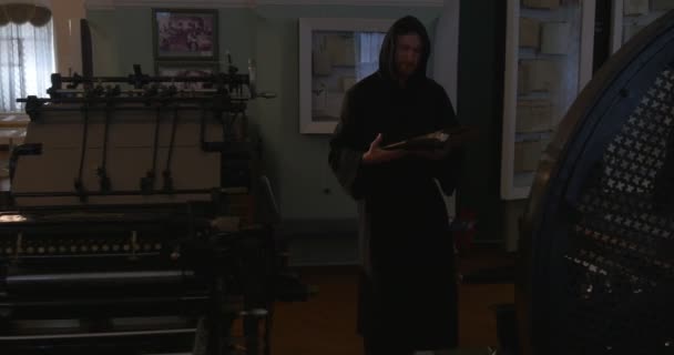 Ο μοναχός περπατάει με το βιβλίο από το δωμάτιο με καφέ ξύλινο πάτωμα, έπιπλα, παράθυρα, εικόνες, αρχαία εργαλεία εκτύπωσης, Μουσείο, ο άνθρωπος διαβάζει — Αρχείο Βίντεο