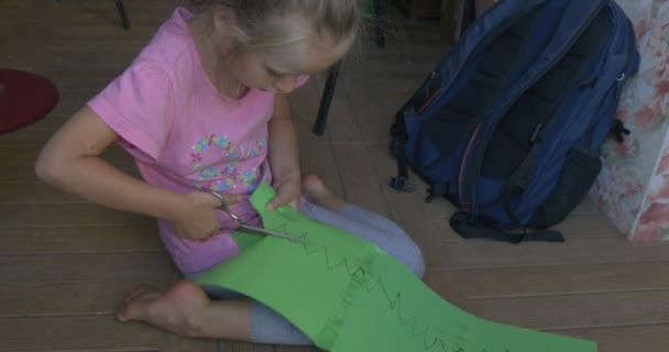 Kleines Mädchen in oinfarbenem T-Shirt sitzt auf dem Boden, Farbpapier vor sich, schneidet die Figur aus grünem Blatt Papier, Rucksack ist in der Nähe — Stockvideo