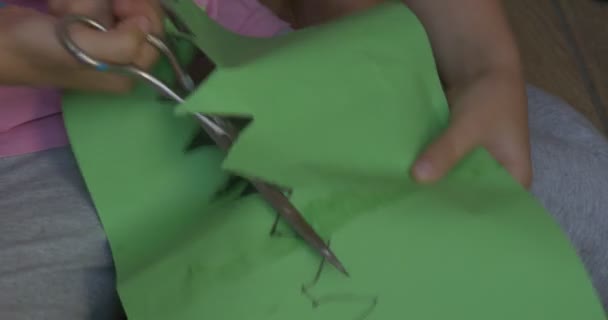 Маленькая девочка в хрустящей футболке сидит на полу и вырезает фигурку короны из зеленого листа бумаги — стоковое видео