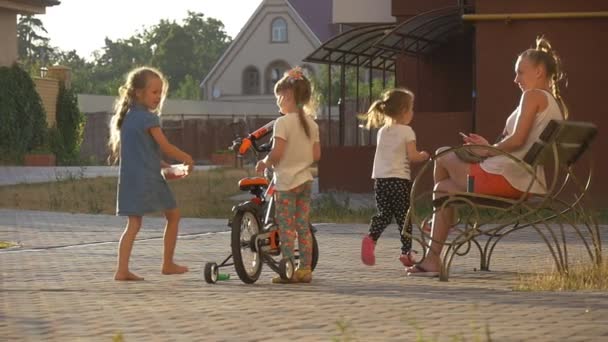 Crianças estão brincando em um parque infantil, no jardim, crianças, menina está segurando a bicicleta, sentado na bicicleta, mamãe está sentada no banco — Vídeo de Stock