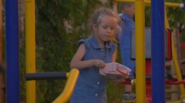 Çocuklar parkta, elinde plastik kutu bir kızla salıncak.