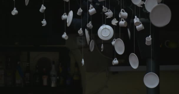 White Mugs, White Saucers, Hunging Under The Ceiling on The Threads, Turning, Bottles on Background, Metal Construction of Ceiling — ストック動画