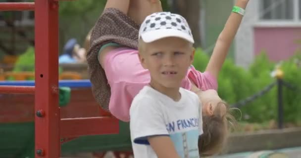 Crianças, crianças estão brincando no parque infantil, menina está pendurada na escada no Playgroung, menino em branco Cap está passando por, Escultura no fundo — Vídeo de Stock