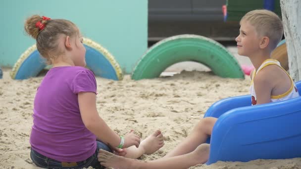 Los niños están jugando, en el parque infantil Sandy, niño y niña en camiseta violeta están sentados en el Blue Chute, Sandy Ground, excavaron neumáticos pintados — Vídeo de stock