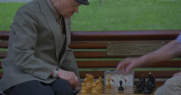 Двое мужчин сидят друг перед другом на скамейке, играют в шахматы, включают шахматные часы, человек указывает на что-то, другой человек делает поворот — стоковое видео