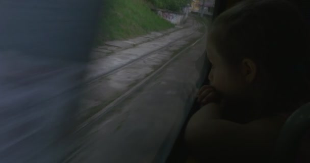 Bambina bionda in camicia rosa è seduto alla finestra in autobus, tram, filobus, guardando Lviv, tram accanto al bus, muro con pianta strisciante — Video Stock