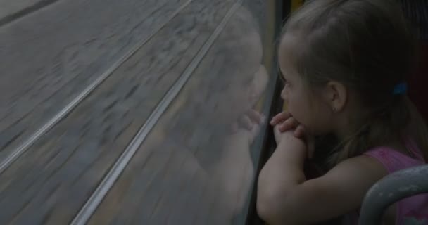 Petite fille blonde en chemise rose est assise à la fenêtre dans le bus, tramway, trolleybus, regarder, Lviv, Fille sur un siège, chemin de fer derrière la fenêtre — Video