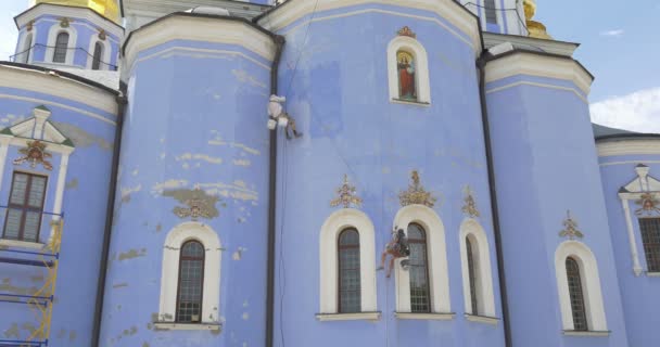 Dos restauradores, hombres, escaladores industriales, distantes, sentados en un asiento sosteniendo una cuerda, catedral de San Miguel, reparación, cúpulas doradas, cielo azul — Vídeo de stock