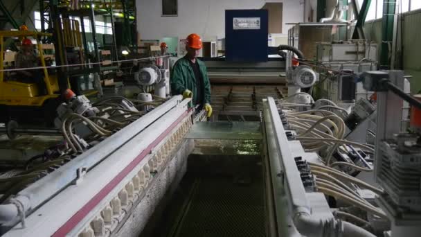 El trabajador está lavando las hojas de vidrio, el trabajador está sentado en un cargador, los trabajadores están en cascos de seguridad naranja, máquina para biselado, producción de vidrio — Vídeos de Stock