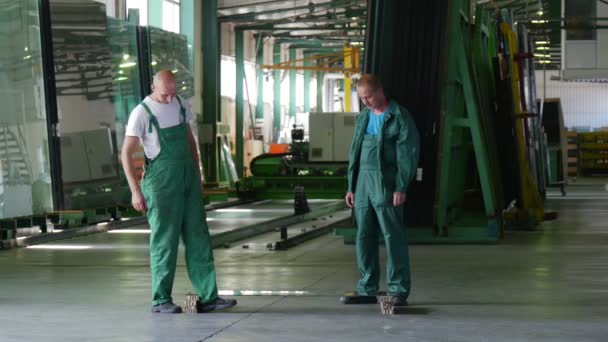 Dos trabajadores en uniforme verde, botas, están de pie en la hoja de vidrio, hoja está acostado horizontalmente en dos bloques, antes de la prueba, prueba de un vaso — Vídeo de stock