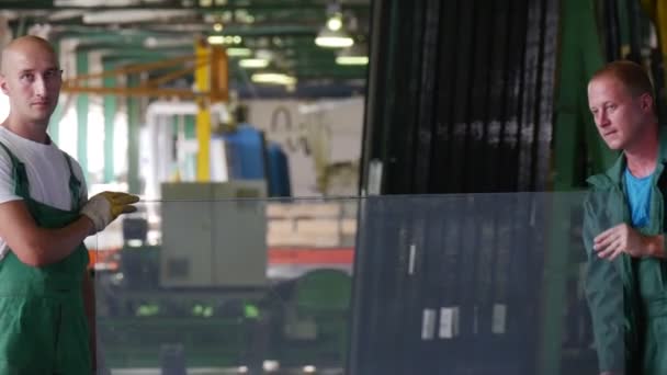Два рабочих в зеленой форме стоят, держа в руках лист стекла, положить лист вниз, испытания пуленепробиваемого стекла, закаленного стекла, стекольный завод — стоковое видео
