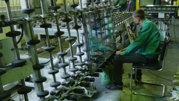 El trabajador con uniforme verde está sentado en la máquina, transportador, llega la hoja de vidrio, el trabajador mira el vidrio, la máquina está girando la hoja horizontalmente — Vídeos de Stock