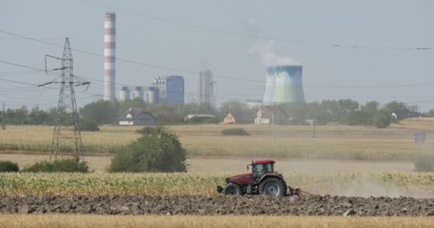 Tractor wordt gedreven door de Fiels landschap droge stro hoogspannings toren draden buizen van de fabriek rook van buizen huizen op een horizon bomen blauwe hemel — Stockvideo