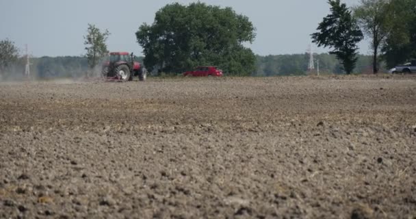 El tractor está soplando distantemente el polvo volador del suelo negro del campo detrás de la carretera del tractor a lo largo de los coches del campo en los árboles de la carretera a lo largo de la carretera — Vídeo de stock
