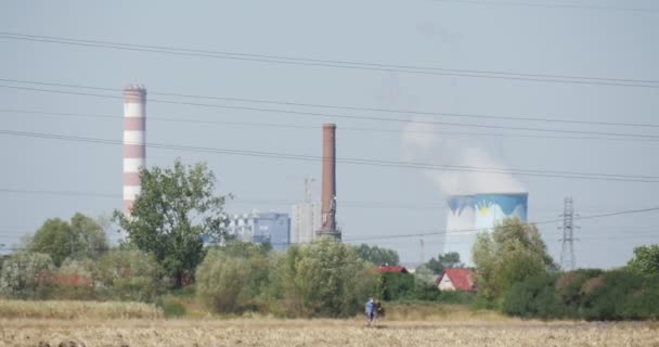 Két az emberek sétálnak a mező csöveket a gyári füst cső vezetékek fák és bokrok házak a láthatáron — Stock videók