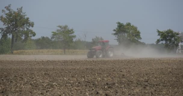 Traktor pflügt das Feld vorbei an Kamera pflügt das Feld lang die Straße fliegenden Staub Autos vorbei an Straßenbäumen neben der Straße — Stockvideo