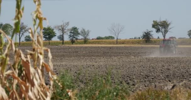 Трактор дистанционно вспахивает поле черной почвы, летящую пыль за тракторной дорогой вдоль полевых автомобилей на дорогах кукурузных стеблей на переднем плане — стоковое видео