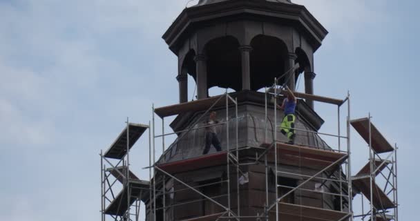 Dos trabajadores en un andamio montan un andamio en una torre alrededor de la torre de la catedral Iglesia Cielo azul Nubes blancas Árboles verdes — Vídeos de Stock