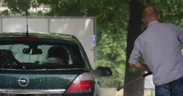 Homem com mangueira lava seu verde escuro Opel Hatchback no Carwash Green Trees Summer Day ao ar livre — Vídeo de Stock