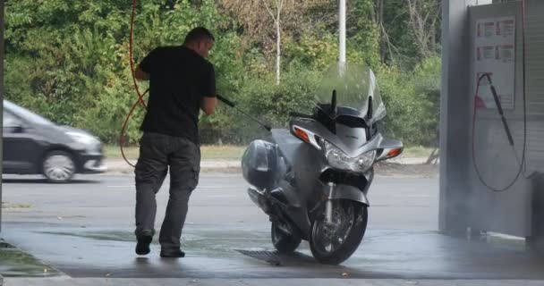 Man Washes His Silver Motorbike At The Carwash Two Bicyclers Pass By Cars Go By Paved Road Behind The Carwash Wet Asfalto Árvores Verdes Dia de Verão — Vídeo de Stock