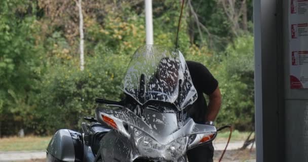 Człowiek z pędzlem ostrożnie myje jego srebrny motocykl na Carwash przedniej szyby w Suds samochód przechodzi przez letni dzień odkryty — Wideo stockowe