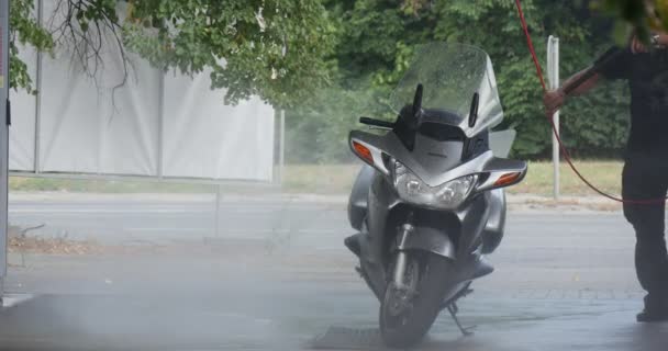 ホースパイプを手に持った男は、舗装された道路夏の日の屋外で行く背景車の洗車で慎重に彼の銀のバイクを洗います — ストック動画