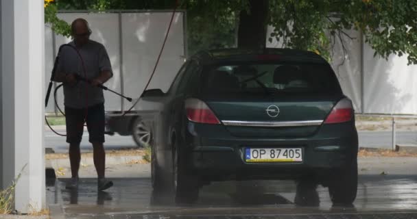 Mann an der Waschanlage wäscht seinen dunkelgrünen Opel Fließheck auf dem Hintergrund verschiedene Autos fahren durch asphaltierte Straße grünen Baum nassen Asphalt Sommertag im Freien — Stockvideo