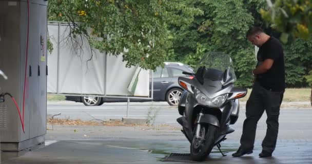 Muž stojí poblíž jeho stříbrné motocyklu na myčka bere čistící Detergent a omývá jeho motocykl mokré asfaltové silnici zelené stromy letní den — Stock video