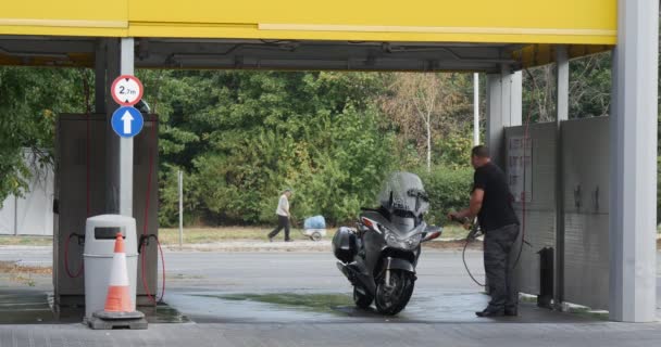 Человек с кистью стирает свой серебряный мотоцикл на автомойке за автомойкой, едет по асфальтированной дороге, а мужчина идет с корзиной Летний день на открытом воздухе — стоковое видео