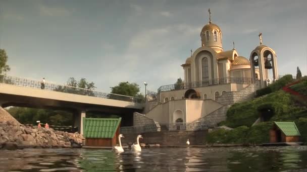 Люди идут по мосту через пруд недалеко от церкви в Донецке Лебеди на пруду Лебединые дома церковь Рядом с металлургическим заводом — стоковое видео