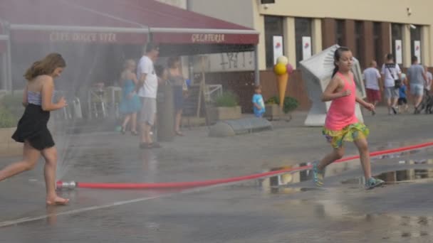 Happy Girls Teens Runs Under Water Splashes en la calle en un caluroso día de verano que hizo que una manguera de agua dañada, cayera en un reposapiés. Cerca con un café adultos y un niño los observa con placer . — Vídeos de Stock