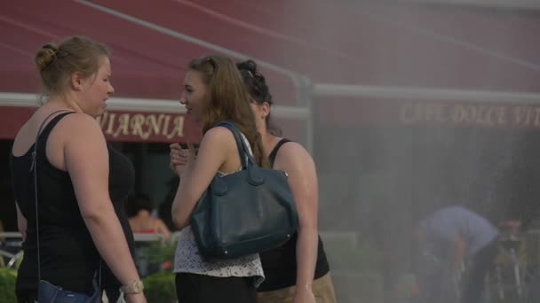 Троє дівчаток середнього віку переговори про добре на вулиці недалеко від кафе. На задньому плані самостійно зроблений фонтан крапель води з пошкоджених пожежних шлангів, що бризки вгору. — стокове відео
