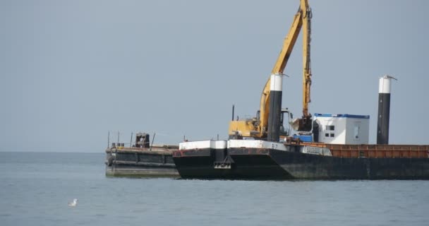 Excavadora amarilla de trabajo en la barcaza La elevación de la creación del suelo de la presa Groynes Obras de construcción en el mar Barco de velocidad roja flota en el mar Día de verano — Vídeos de Stock