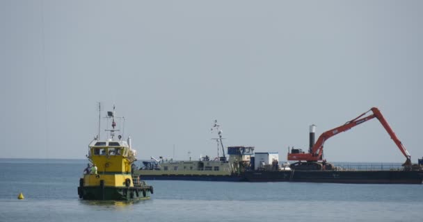 Big pråm med grävmaskin på tavlan gul-grön båt flyter på havet skapandet av dammen groynes byggnad arbetar på havet människor på havsstranden sommardag — Stockvideo