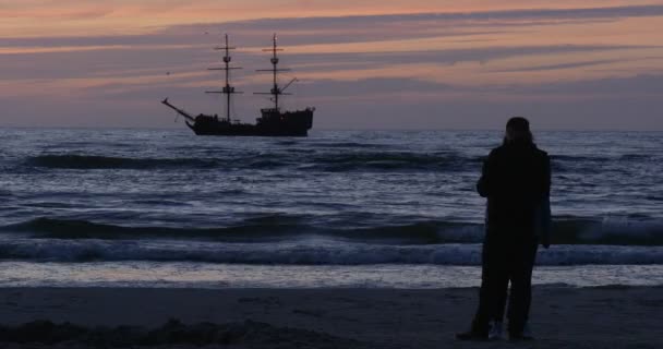 Paar Mann und Frau stehen an der Küste am Meer bewegliches Schiff auf Hintergrund rosa Himmel Wellen Wolken Abend Sonnenuntergang im Freien — Stockvideo