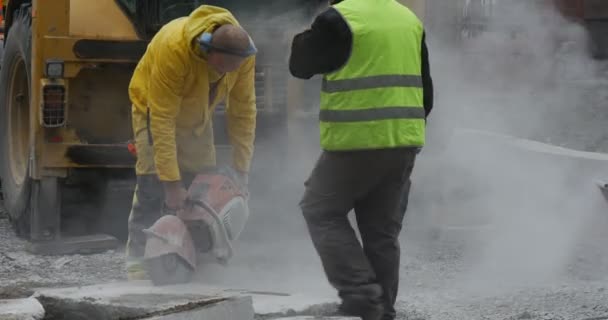 Man werknemer in gele werkkleding is het slijpen van de blokken van beton doorslijpen machine vliegende stof weg reparatie een andere werknemer nadert wuivende hand — Stockvideo