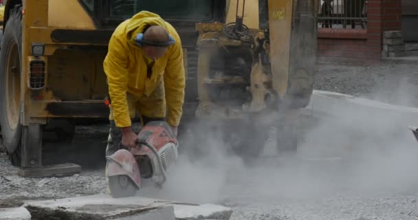 Man werknemer in gele werkkleding is het slijpen van de blokken van beton doorslijpen machine close-up gele graafmachine vliegende stof weg reparatie — Stockvideo