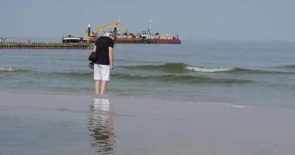 Człowiek z butami w Hand spacery boso na morzu brzegu i Wets jego stopy w morzu wody Surf Breakwater w odległości budowa Barka na morzu — Wideo stockowe
