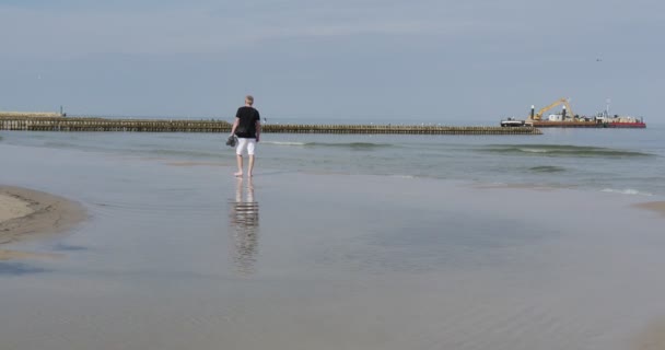Человек в ботинках в руках ходит босиком по берегу моря и мочит ноги в волнорезы на морском волне на расстоянии строительной баржи у моря — стоковое видео