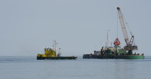 Építőipari uszály daru az igazgatóság bekapcsolja a víz sárga-zöld csónak áll közelében létrehozása Dam groynes építőipari művek a tengeren — Stock videók