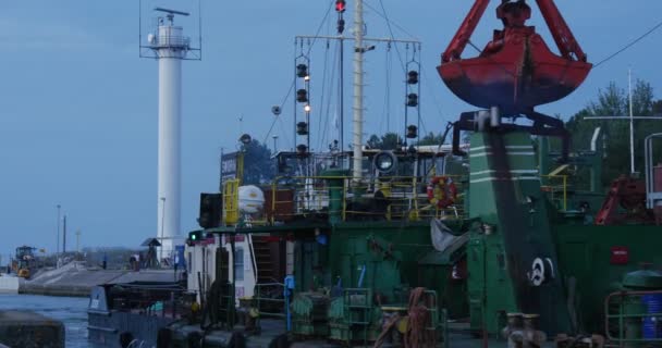 Grön konstruktion fartygs pråm med kran på brädet simmar in i hamn arbetaren i orange arbetsdräkt på tavlan folk går på kajen skymning — Stockvideo