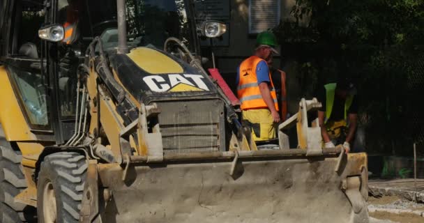 Homem motorista em vestuário de trabalho na escavadeira amarela CAT é trabalhadores permanentes em roupas de trabalho amarelas e capacetes estão trabalhando reparação rodoviária pavimentação da rua Road City — Vídeo de Stock