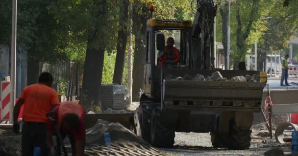 Adam sürücüsüdür iş giysisi sarı Ekskavatör kabin içinde yaklaşan blok işçilerin yığın gerekli ön plan yeşil ağaçlar şehir sokak üzerinde kaldırır — Stok video