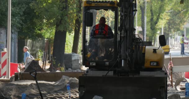 Man arbetare i gula arbetskläder sitter i kabinen på grävmaskin får ut av stuga mångfald Street gul grävmaskin Road reparation byggnader gröna träd — Stockvideo